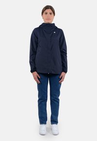 Giacca leggera blu navy con zip frontale, cappuccio e polsini elasticizzati, abbinata a jeans blu e sneakers bianche. Design semplice e urbano.