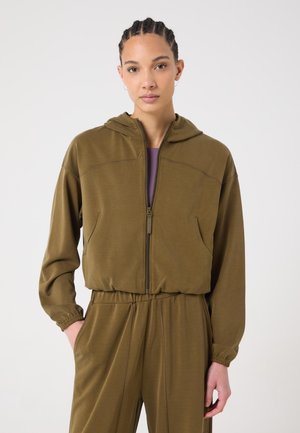 Frau mit geflochtenem Haar, die eine olivgrüne Kapuzenjacke mit Reißverschluss und passende Hosen mit elastischem Bund trägt, die Hände in den Taschen.