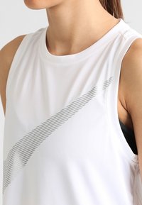 Acercamiento de una persona que lleva una camiseta deportiva sin mangas blanca con un gran logotipo de diseño en forma de swoosh de rayas diagonales grises.