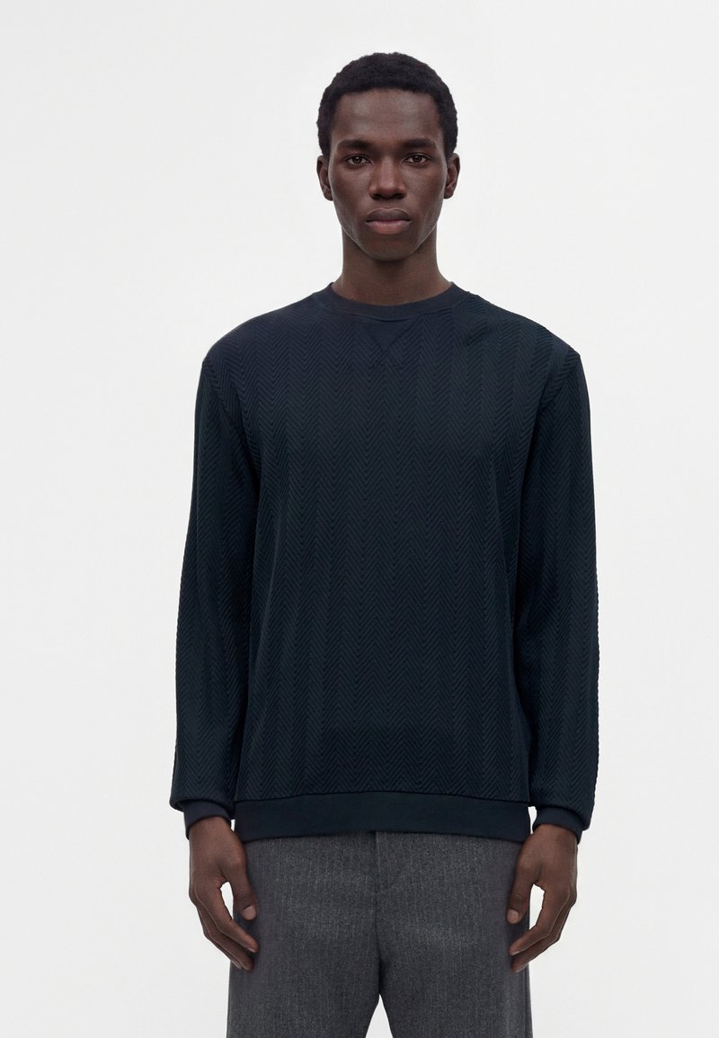Jeune homme vêtu d'un pull à manches longues sombre à motif et d'un pantalon gris, debout devant un fond clair uni.