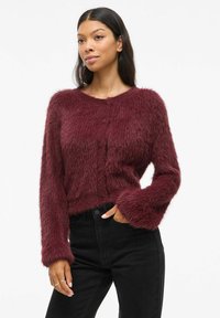 Cardigan bordeaux en peluche avec des manches longues et un col rond. Présente une fermeture à boutons et des poignets côtelés. Associé avec un jean noir.