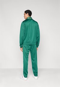 Πράσινη φόρμα από λείο, γυαλιστερό υλικό, που περιλαμβάνει μπουφάν με φερμουάρ και ψηλό γιακά με ελαστικές μανσέτες, συνδυασμένη με παντελόνια στο ίδιο σχέδιο.