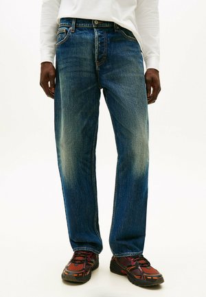 Blauwe spijkerbroek met een rechte pijp, subtiele vervaging, vijfzakkenstijl en een klassieke ceintuurlussentaille. G gedragen met sneakers met rode accenten.