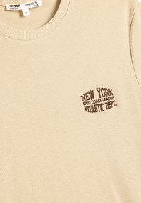 Beige ribbet t-shirt med rund halsudskæring. Har mørkebrune broderede tekster: "New York East Coast League Athletic Dept." på brystet.