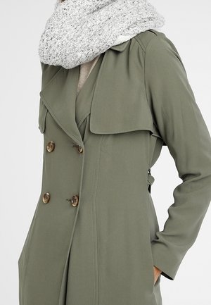Femme portant un trench-coat vert olive à double boutonnage avec des boutons en bois et une grosse écharpe tricotée gris clair.