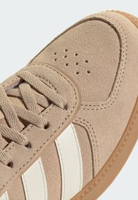 Sneaker i beige mocka med vita läderband, rundad tå, texturerad yta och snörning. Har perforeringar för ventilation.