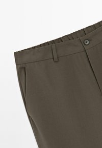Mörk olivgrön byxor med elastisk midja, bälteshällor, sidoficka och svart knappstängning på en vit bakgrund.