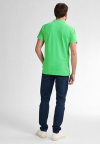 Tricou polo verde cu mâneci scurte, purtat cu blugi negri și adidași de culoare deschisă, văzut din spate. Design simplu și croială lejeră.