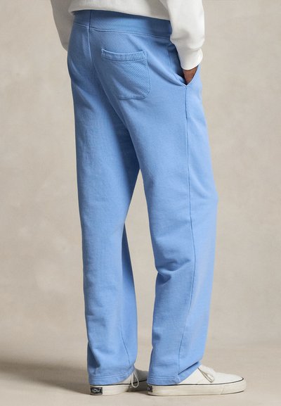 Inimene, kes kannab helesiniseid dressipükse ja valgeid tossusid, käed taskus, seisab neutraalse taustaga.