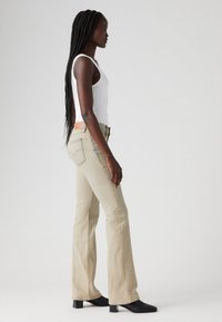 Jean beige évasé avec une couture contrastante, design à cinq poches et une patte en cuir brun à l'arrière. Porté avec un débardeur blanc et des bottes noires.