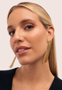 Pendientes de oro con borlas de cadena que caen, cuelgan de las orejas del modelo, con una parte superior metálica suave. El modelo tiene el cabello lacio y claro.