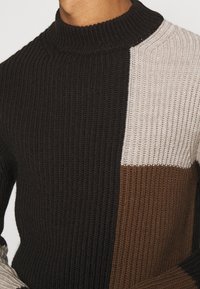 Stickad polotröja i svarta, beige och bruna nyanser. Har vertikal ribbad textur och en kontrasterande färgblockdesign.