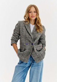 Žena s dlouhými blond vlasy, oblečená v šedém kostkovaném sakou s fialovými knoflíky a volnými modrými džíny, s rukama v kapsách, na neutrálním pozadí.