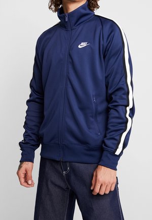 Mężczyzna ubrany w granatową kurtkę sportową Nike z białymi i czarnymi paskami na rękawach oraz w ciemnoniebieskie dżinsy, stojący na tle szarego tła.