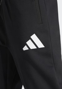 Černé sportovní kalhoty se velkým bílým logem Adidas a třemi diagonálními pruhy na levé straně, vyrobené z hladkého a pružného materiálu.