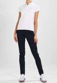 Vit polo-shirt med krage och liten logotyp, kombinerad med marinblå slim-fit byxor. Modell bär vita sneakers med röda accenter.