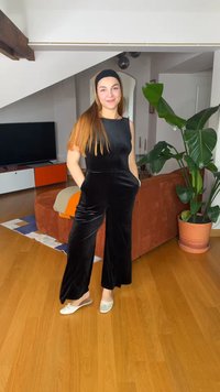 Νεαρή γυναίκα με μακριά μαλλιά και κορδέλα, που φοράει μαύρη ολόσωμη φόρμα και λευκά παπούτσια, στέκεται χαμογελώντας σε ένα μοντέρνο σαλόνι με φυτά και ξύλινο δάπεδο.