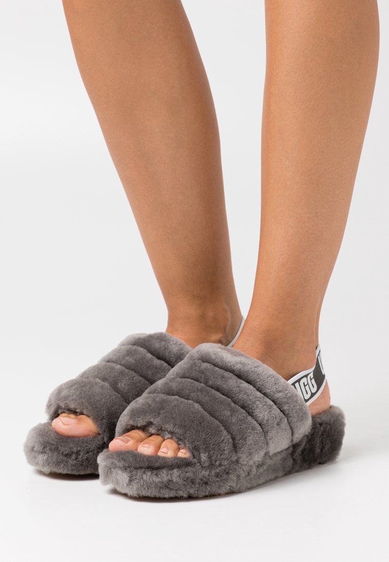 Pieds portant des chaussons gris duveteux ouverts à l'avant avec une sangle élastique noire et blanche de marque au talon, posés sur une surface blanche.