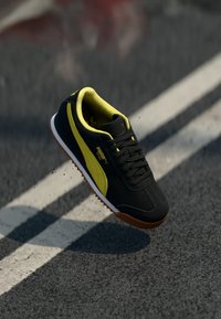 Chaussure de sport noire avec accents jaunes, tige texturée et semelle en caoutchouc. Design à lacets avec des pointes de semelle extérieures visibles.