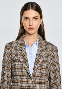 Hnědý kostkovaný blazer s klopovým límcem, který má zapínání na jeden knoflík a texturovanou tkaninu. Nosený přes světle modrou košili s límečkem.