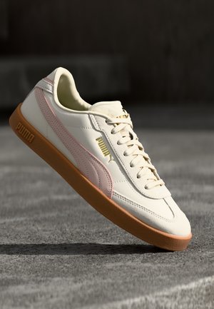 Biały sneaker Puma z różowym paskiem i gumową podeszwą unoszący się nad teksturowaną, szarą powierzchnią w ciemnym otoczeniu.