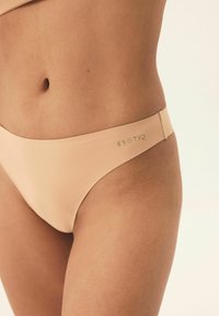 Beiger Bikini-Unterteil mit glatter Textur, niedrig geschnitten und minimalen Nähten. Mit dem Logo "ESOTIQ" in einem kontrastierenden Farbton am Bund.