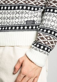 Neulottu villapaita, jossa on valkoisen, ruskean ja mustan yhdistelmä, geometrisia kuvioita ja teksturoitu resorivahvistus helmassa. Tarralappu näkyvissä sivussa.