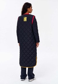 Žena s tmavými vlasy, oblečená v dlouhém prošívaném černém kabátu s červeným a žlutým lemováním, tmavých kalhotách a bílých teniskách, stojící bokem.