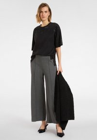 Czarna bluzka z cekinami na ramionach, w połączeniu z szerokimi szarymi spodniami i strukturalną czarną kurtką. Modelka nosi czarne szpilki.