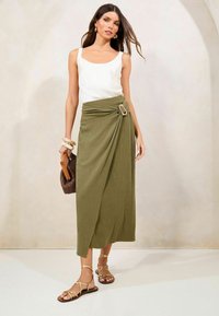 Valge tops, oliivroheline ümbermooditud seelik, millel on klambriga detail, koos rihmadega sandaalide ja kootud käekotiga. Sujuv kanga tekstuur ning minimalistlik disain.