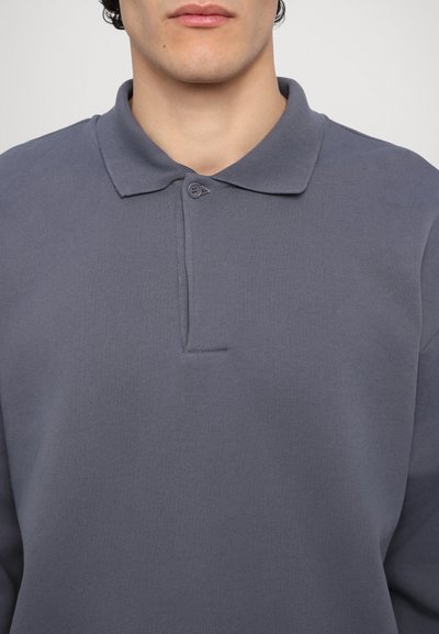 Šedé polo tričko vyrobené z texturované látky, s knoflíkovou légou a klasickým límcem. Design je jednoduchý a nenápadný.