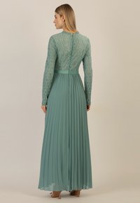 Abito lungo plissettato in teal, con corpo in pizzo, maniche lunghe e scollatura a coste. Chiusura con cerniera sul retro visibile.