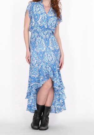 Femme en robe bleue fleurie à ourlet évasé, associée à des bottes noires à boucle mi-mollet, debout contre un fond blanc.