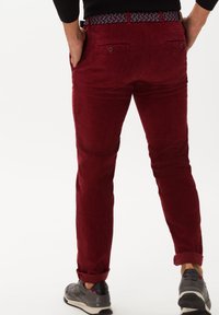 Pantalons en velours côtelé rouge avec une coupe slim, présentant des ourlets retroussés, des poches arrière avec des fermetures à bouton et une ceinture à motifs. Portés avec des baskets grises.
