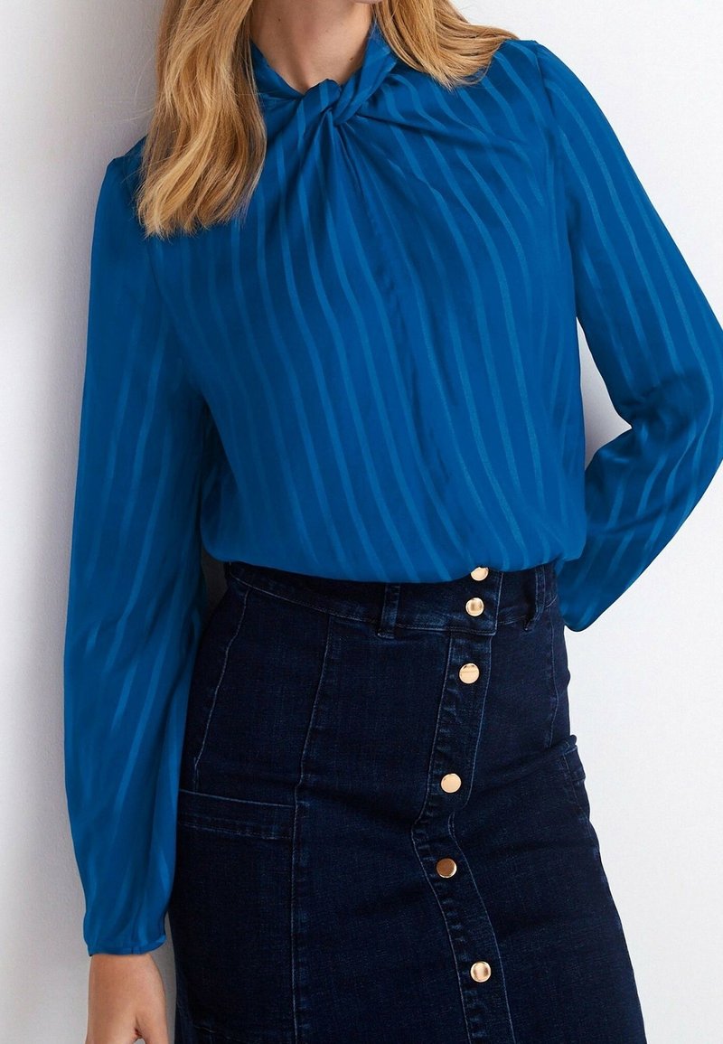 Femme portant un chemisier bleu à manches longues avec de fines rayures et une jupe en jean foncé taille haute avec des boutons dorés sur le devant.
