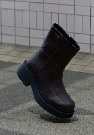 Bottine en cuir marron foncé avec une semelle noire épaisse posée sur un sol pavé devant un mur carrelé.