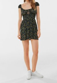 Nainen, joka on pukeutunut lyhyeen mustaan kukkamekkoon, jossa on puffihihat, ja valkoisiin tennareihin, seisoo yksinkertaista valkoista taustaa vasten.