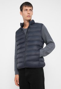 Junger Mann in einer marineblauen Steppweste über einem grauen Strickpullover und schwarzen Hosen, der vor einem schlichten weißen Hintergrund steht.