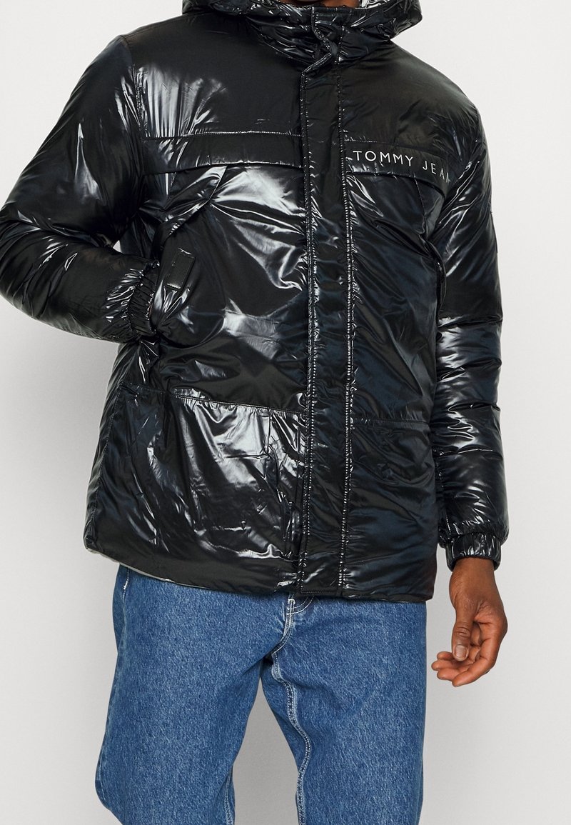 Homme portant une doudoune noire brillante à capuche avec le logo "Tommy Jeans", associée à un jean en denim bleu, mains partiellement visibles.
