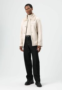 Beige jas met een zachte kraag, gedragen over een lichte coltrui. Zwarte jeans met wijde pijpen en zichtbare naden. Zwarte laarzen maken de look compleet.