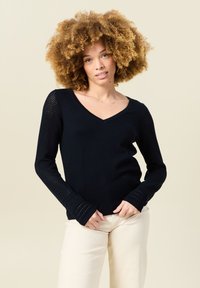 Svart stickad tröja med V-ringning, med texturerade meshärmar och figursydda muddar. Bärs med ljusa byxor.