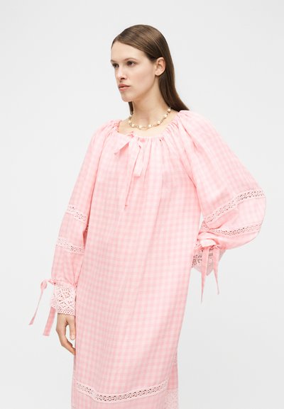 Γυναίκα που φορά χαλαρό ροζ φόρεμα γκινγκχαμ με φουσκωτά μανίκια, διακόσμηση με δαντέλα και κορδέλες, συνδυασμένο με κολιέ από πέρλες.