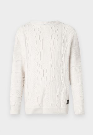 Valkoinen kaarrokepaita, jossa on monimutkaisia tekstuurikuvioita, resorikainalot, helma ja pyöreä pääntie, esillä yksinkertaisella taustalla.