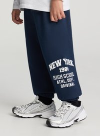 Granatowe spodnie dresowe z białym nadrukiem „NEW YORK 1984 HIGH SCHOOL ATHL. DEPT. ORIGINAL”, zestawione ze srebrnymi butami sportowymi z siateczkowymi detalami.