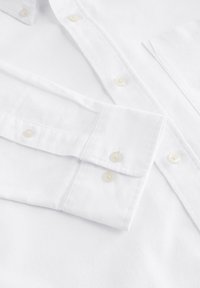 Camicia bianca in cotone con polsini a bottoni, con bottoni dello stesso colore. Tessuto liscio, design del colletto classico e dettaglio di taschino singolo.