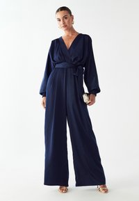 Marineblauwe satijnen jumpsuit met wijde benen, diepe V-hals, ceintuur in de taille en lange mouwen; gecombineerd met een metallic clutch en gespenhakken.