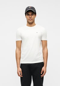 Άνδρας με λευκό κοντομάνικο μπλουζάκι, σκούρο παντελόνι και σκούφο μπέιζμπολ, που στέκεται μπροστά σε απλό φωτεινό φόντο.