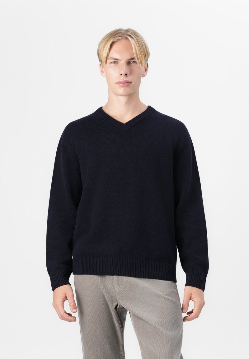 Pull en V marine avec des poignets et un ourlet côtelés, fabriqué en tissu doux, associé à un pantalon en velours côtelé de couleur claire. Design simple et classique.