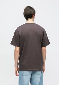 Νεαρό άτομο με κοντά σκούρα μαλλιά φοράει φαρδιά σκούρη γκρι μπλούζα και ανοιχτόχρωμο τζιν, στραμμένο με την πλάτη προς λευκό απλό φόντο.