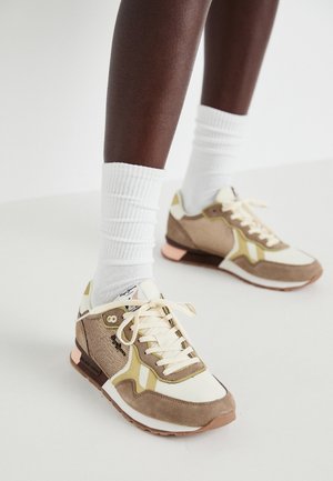Zapatillas deportivas en tonos beige y crema con acentos amarillos, materiales de ante y malla, diseño en capas y suela de color rosa claro, usadas con calcetines blancos.
